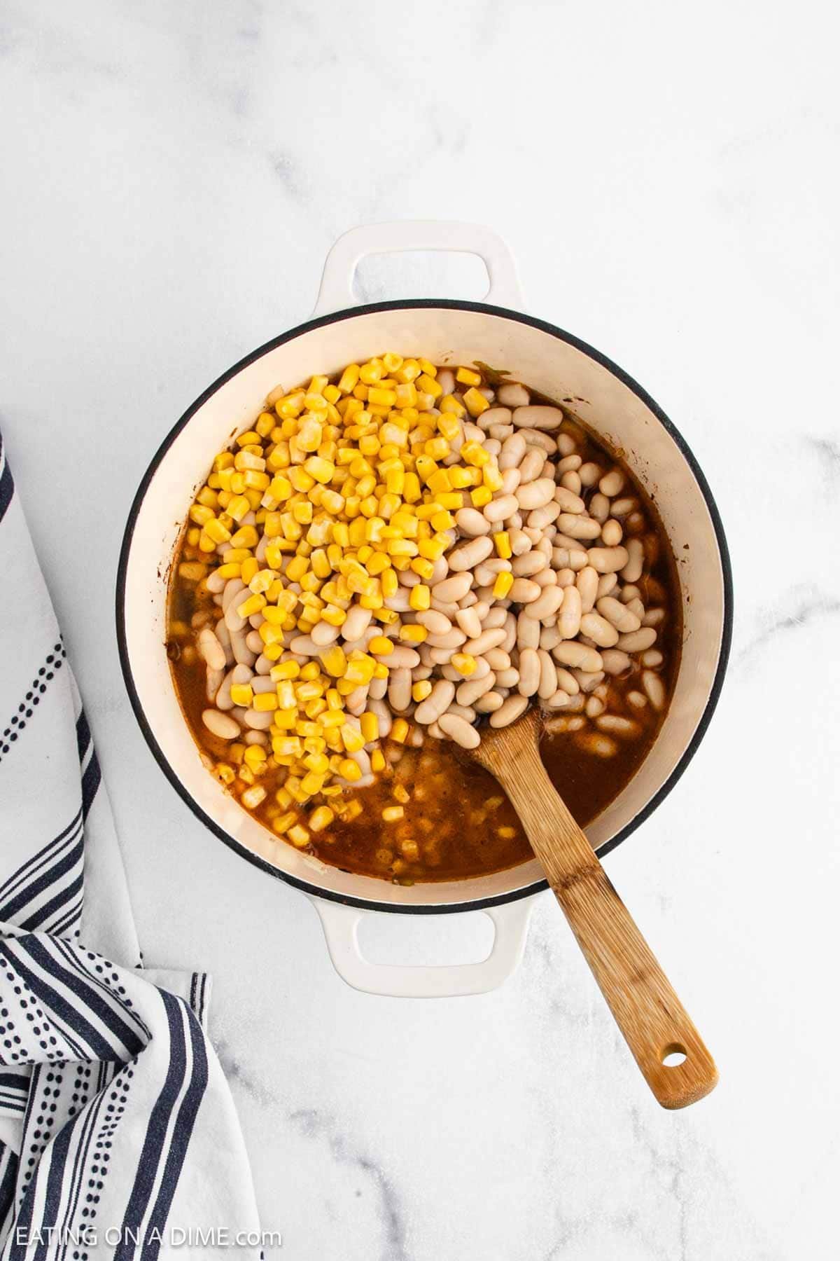 A white pot filled with hearty White Bean Turkey Chili, featuring broth, white beans, and corn, sits on a marble surface. A wooden spoon rests inside the pot, and a striped towel is nearby.