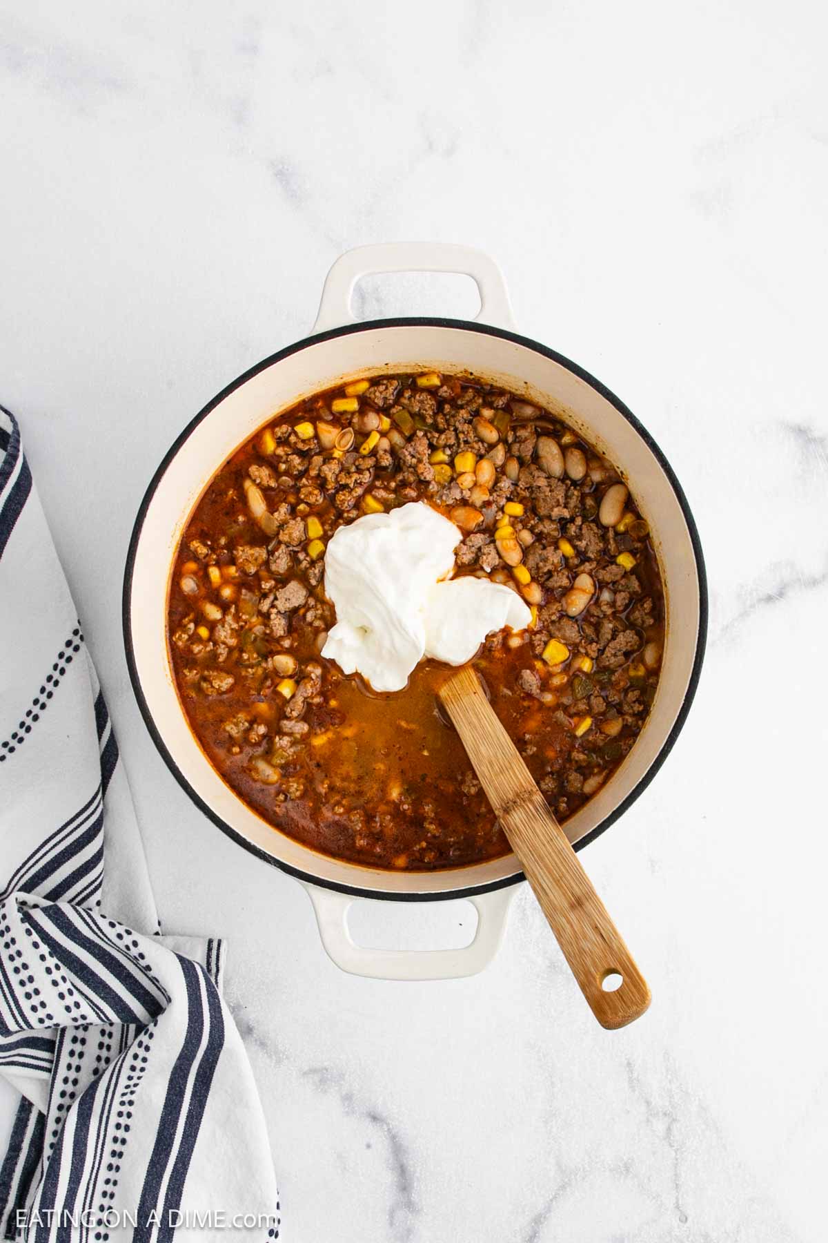 A white pot filled with hearty White Bean Chili topped with a dollop of sour cream. A wooden spoon rests inside the pot, and a striped kitchen towel is placed beside it on a white countertop.