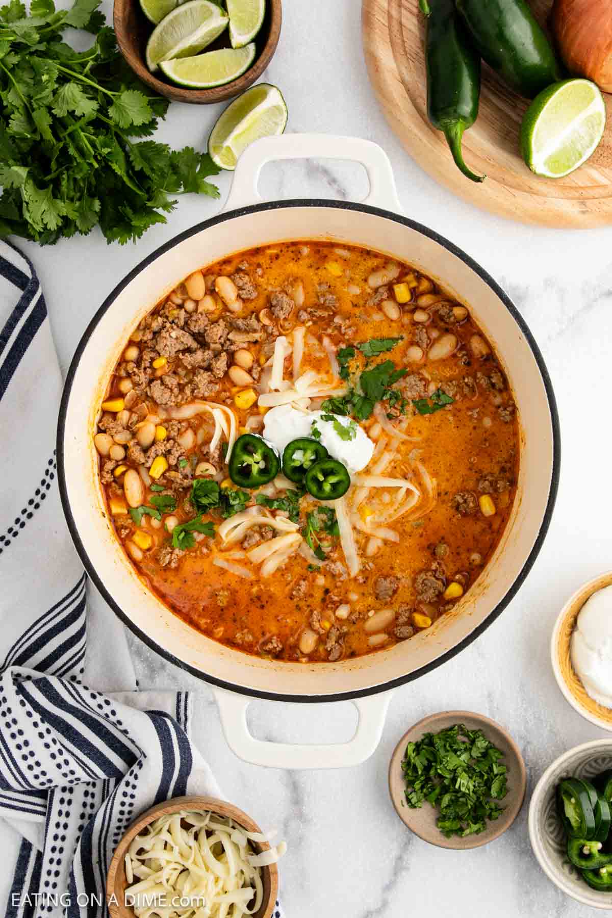 A pot of creamy White Bean Chili soup topped with sour cream, shredded cheese, jalapeño slices, and cilantro, surrounded by bowls of toppings and fresh ingredients like limes, onions, and jalapeños on a marble surface.