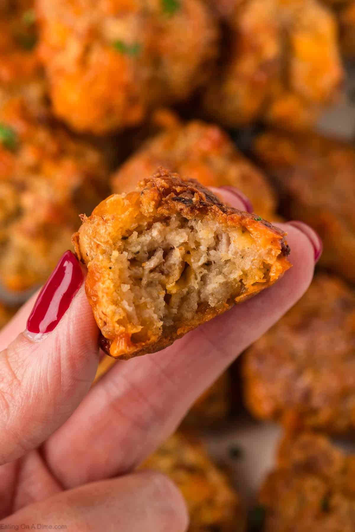 A close-up of a hand with red nail polish holding a bite-sized cream cheese sausage ball, showing its moist, textured inside. More cream cheese sausage balls are blurred in the background.