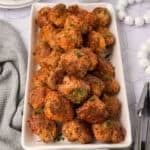 A rectangular white platter filled with golden-brown Cream Cheese Sausage Balls garnished with chopped parsley, placed on a white surface next to a gray textured cloth and a pair of tongs.