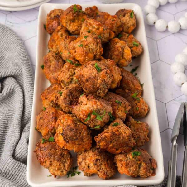 A rectangular white platter filled with golden-brown Cream Cheese Sausage Balls garnished with chopped parsley, placed on a white surface next to a gray textured cloth and a pair of tongs.