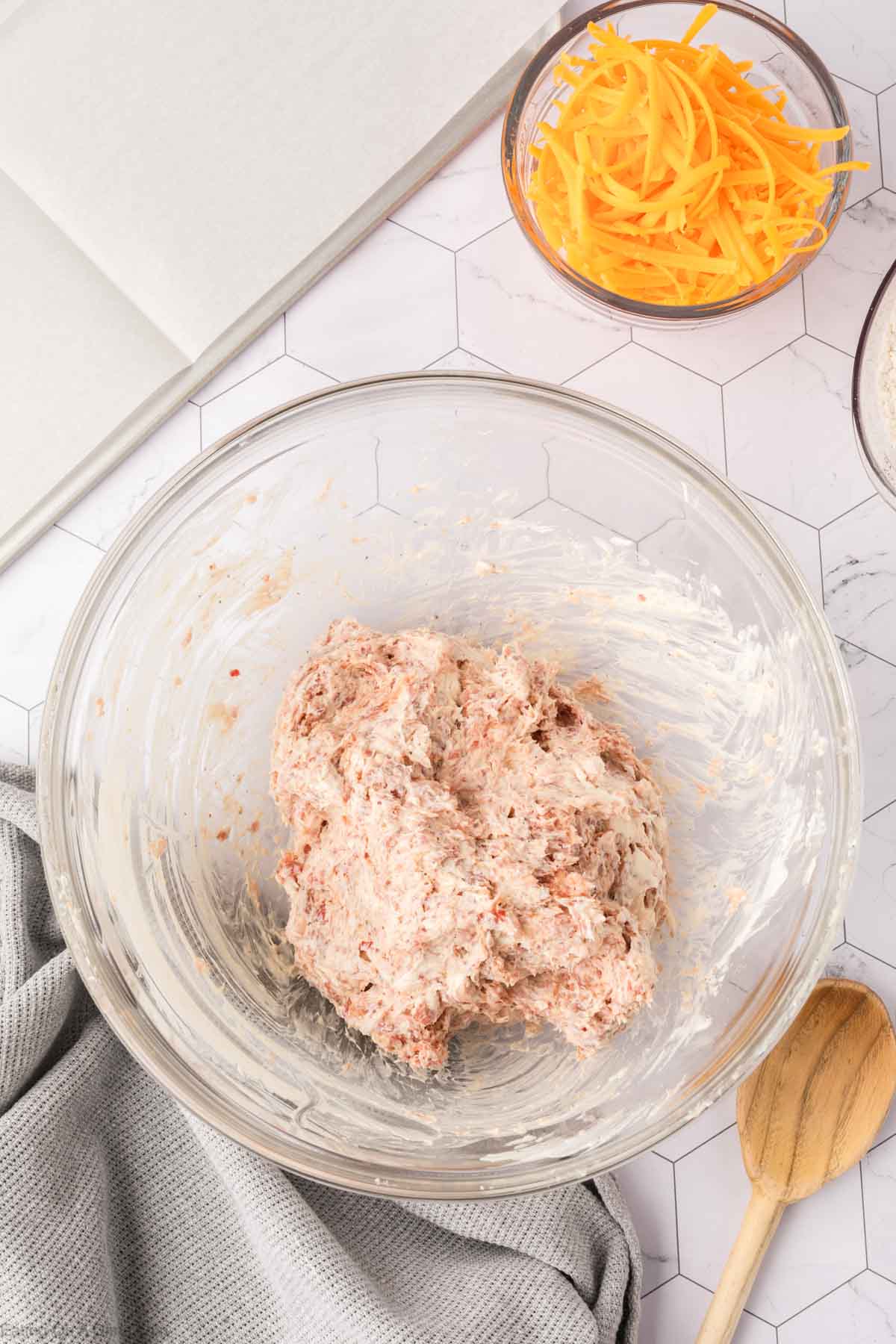 A glass bowl with biscuit dough sits on a counter next to a small bowl of shredded cheddar cheese, perfect for making Cream Cheese Sausage Balls. A wooden spoon, gray towel, and open notebook complete the cozy kitchen scene.