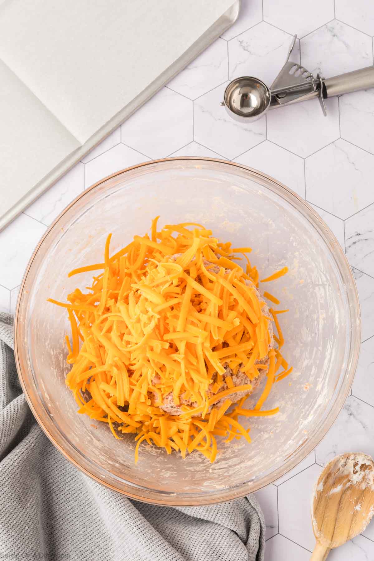A glass mixing bowl filled with shredded cheddar cheese and the base for Cream Cheese Sausage Balls sits on a countertop, surrounded by a wooden spoon, gray towel, open notebook, and an ice cream scoop.