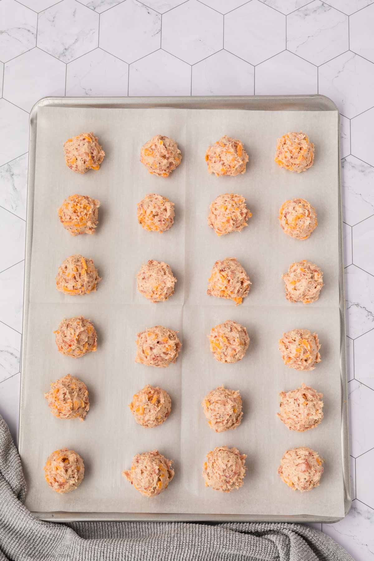 A baking sheet lined with parchment paper holds 20 evenly spaced, round scoops of uncooked cream cheese sausage balls, ready to be baked. The tray sits on a marble surface, perfect for delicious cream cheese appetizers.