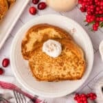 Two slices of Eggnog French Toast on a white plate, topped with whipped cream and a sprinkle of cinnamon. Cranberries, red berries, and a red-striped towel are nearby on a white table.