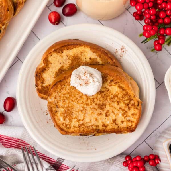 Two slices of Eggnog French Toast on a white plate, topped with whipped cream and a sprinkle of cinnamon. Cranberries, red berries, and a red-striped towel are nearby on a white table.