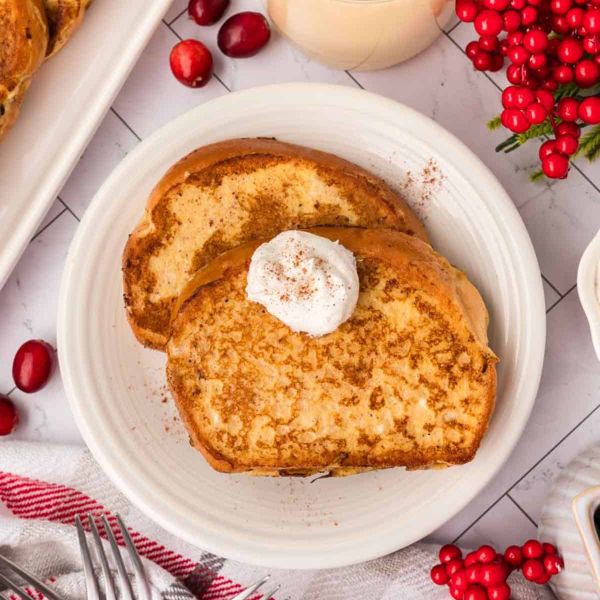 Two slices of Eggnog French Toast on a white plate, topped with whipped cream and a sprinkle of cinnamon. Cranberries, red berries, and a red-striped towel are nearby on a white table.