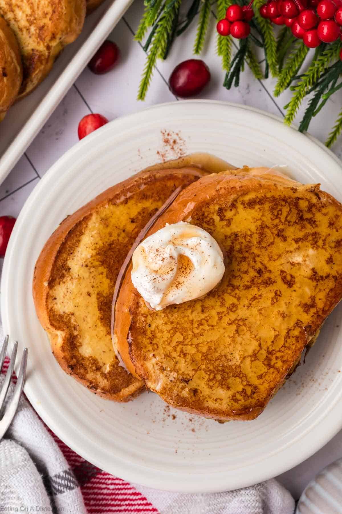 Two slices of golden Eggnog French Toast topped with a dollop of whipped cream and a sprinkle of cinnamon, served on a white plate with holiday decorations and a red-and-white towel nearby.