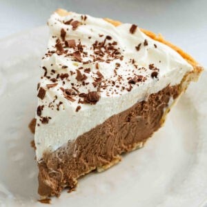 A slice of French Silk Pie with a thick layer of whipped cream on top, sprinkled with chocolate shavings, sits on a white plate.