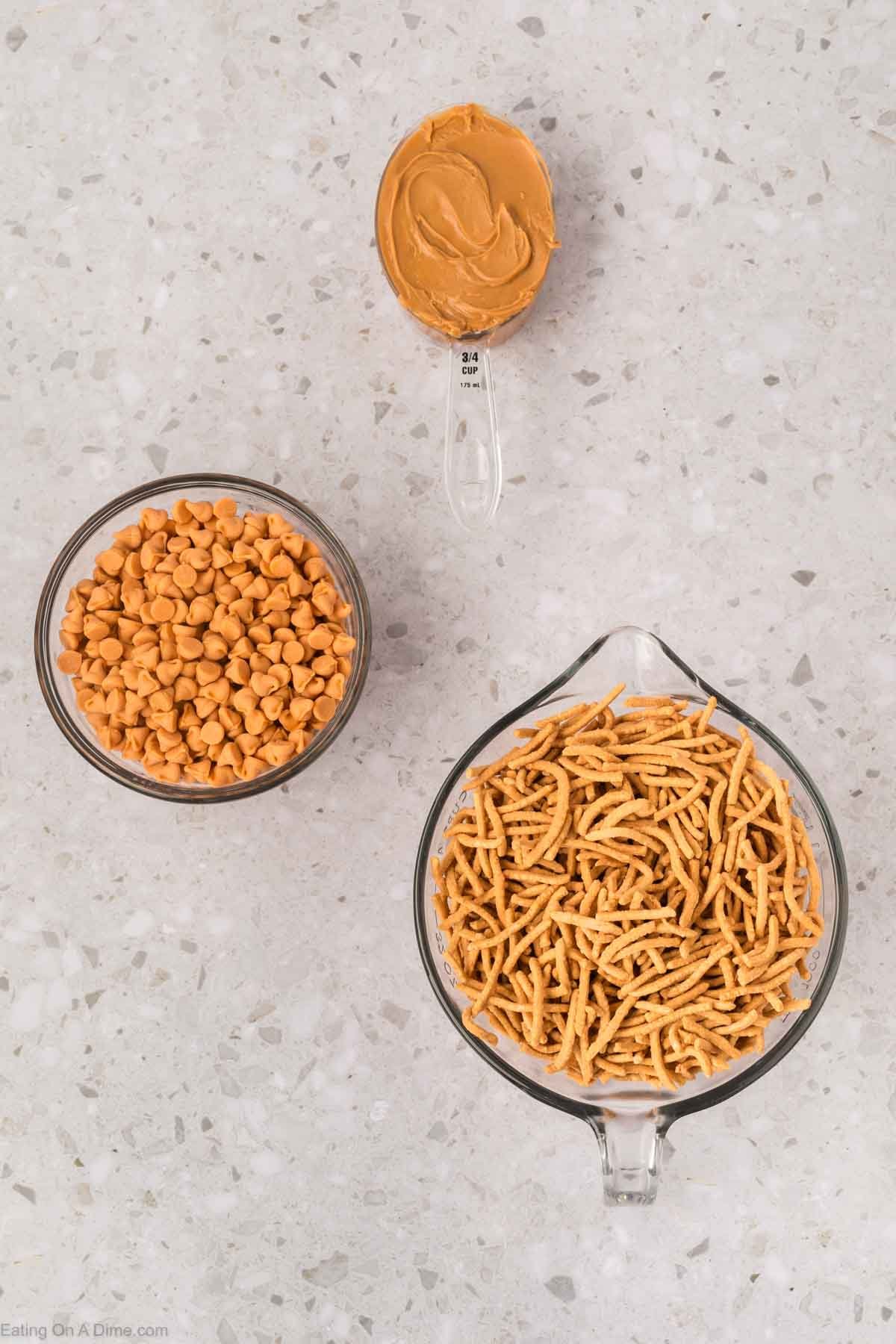 Overhead view of three ingredients for Haystack Cookies on a countertop: a measuring cup of crispy chow mein noodles, a bowl of butterscotch chips, and a tablespoon of creamy peanut butterโperfect for easy no-bake cookies or chocolate treats.