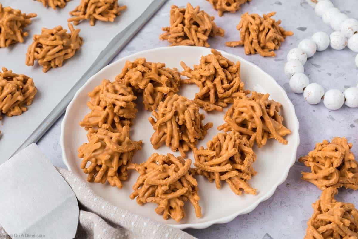 A white plate holds several clusters of haystack cookies, with more no bake cookies arranged on parchment paper in the background. The light brown treats have a lumpy, nest-like appearance, perfect for chocolate haystack lovers.
