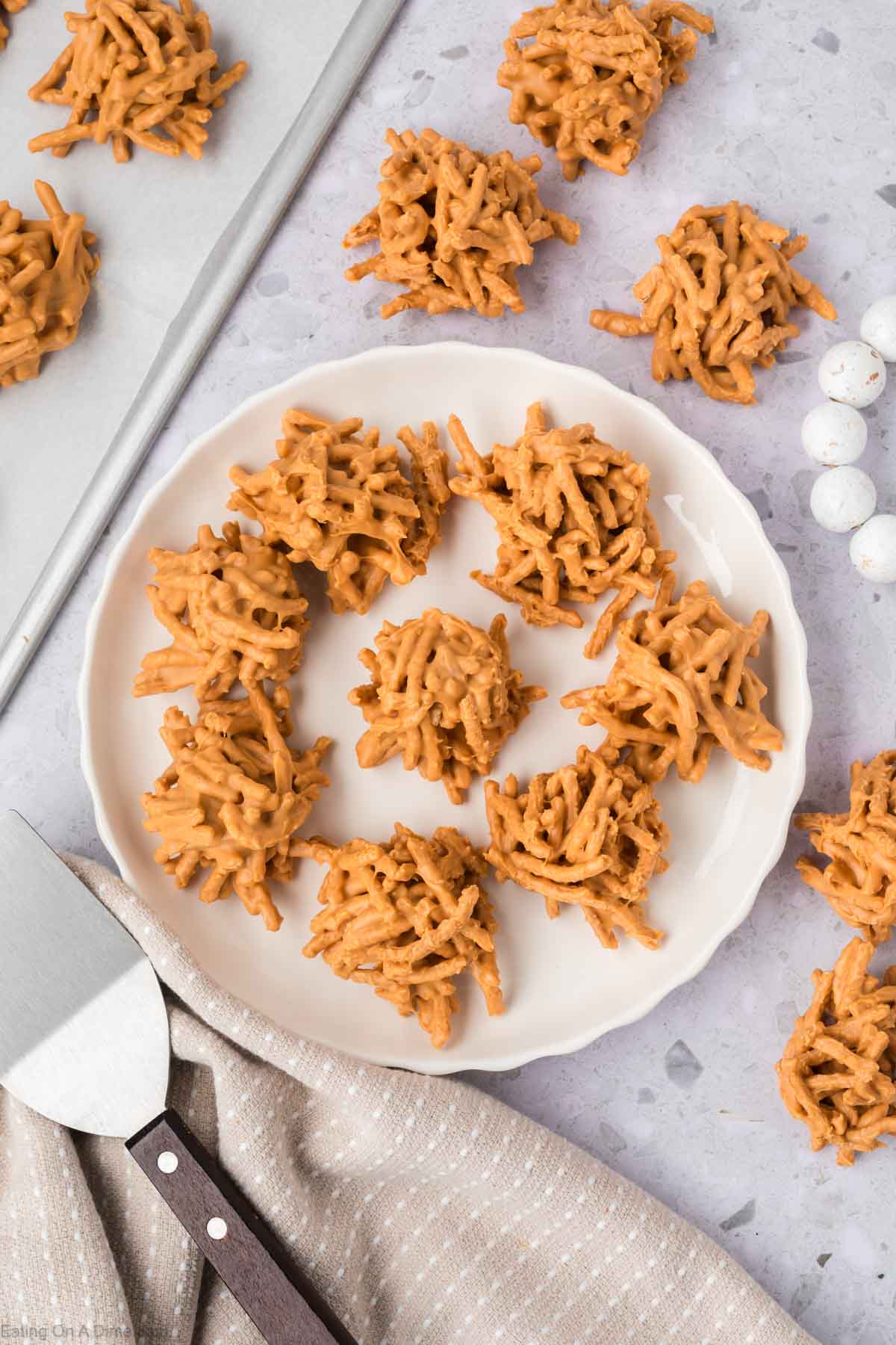 A white plate filled with clusters of Haystack Cookies sits on a light countertop, surrounded by more cookies, a spatula, and a beige kitchen towel.