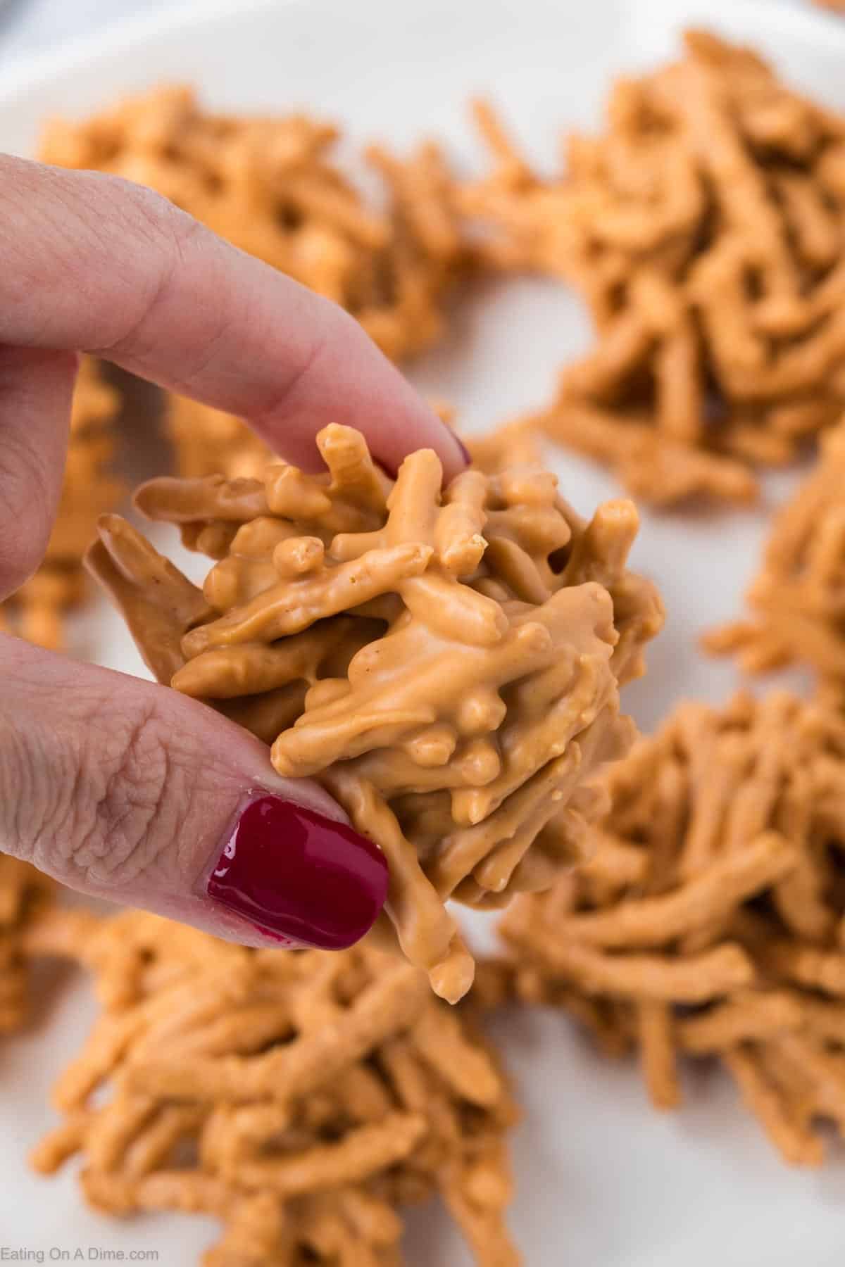 A hand with red nail polish holds a classic Haystack Cookie made of crunchy noodles coated in a creamy peanut butter mixture, with more Haystack Cookies on a white plate in the background.