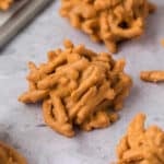 Close-up of a classic Haystack Cookie—an easy cookie recipe made from chow mein noodles coated in a light brown, peanut butter-like mixture. Other no bake cookies are arranged in the background on a light gray surface.
