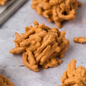 Close-up of a classic Haystack Cookie—an easy cookie recipe made from chow mein noodles coated in a light brown, peanut butter-like mixture. Other no bake cookies are arranged in the background on a light gray surface.