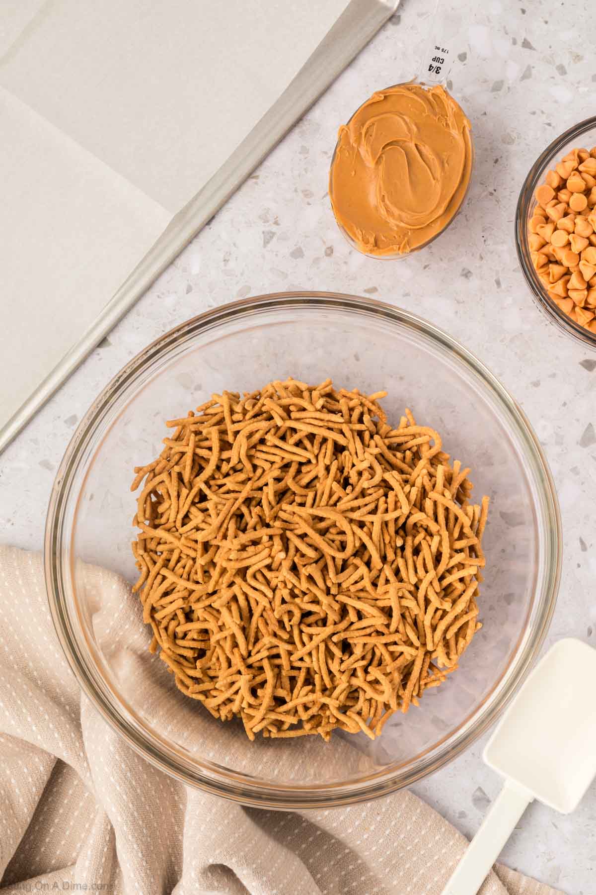 A glass bowl filled with crunchy chow mein noodles is on a countertop next to a cup of creamy peanut butter, butterscotch chips, and baking toolsโclassic ingredients for making irresistible Haystack Cookies.