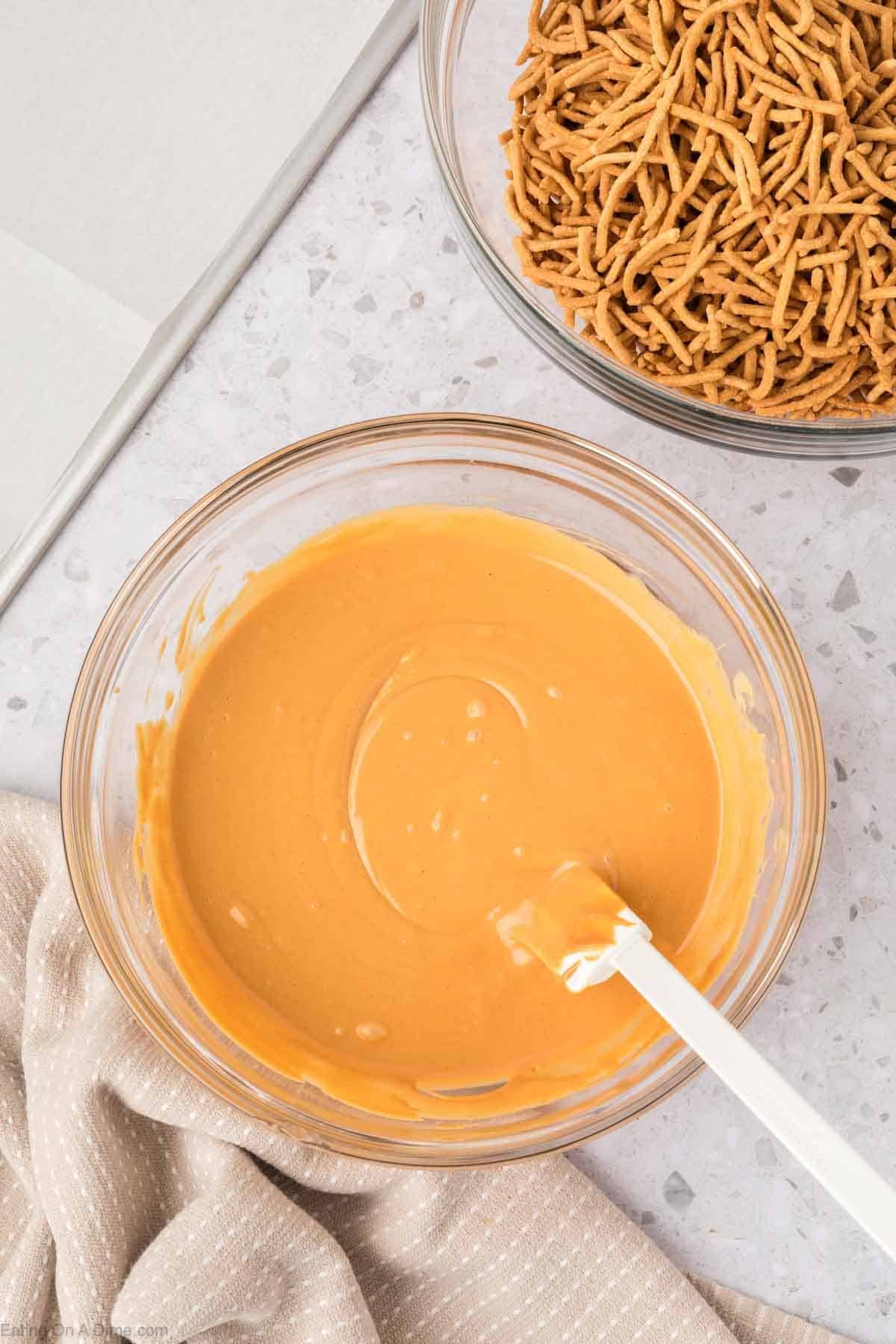 A glass bowl filled with melted butterscotch mixture and a white spatula sits next to a second bowl containing crunchy chow mein noodlesโingredients ready for making classic Haystack Cookies on a light countertop.