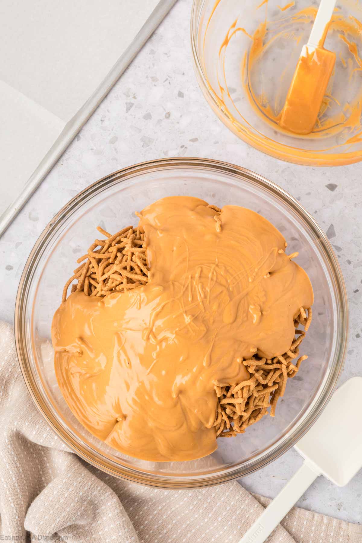 A glass bowl filled with crunchy chow mein noodles topped with melted butterscotch chips resembles classic Haystack Cookies. Beside it are a spatula, a napkin, and another bowl streaked with butterscotch residue, all on a gray countertop.