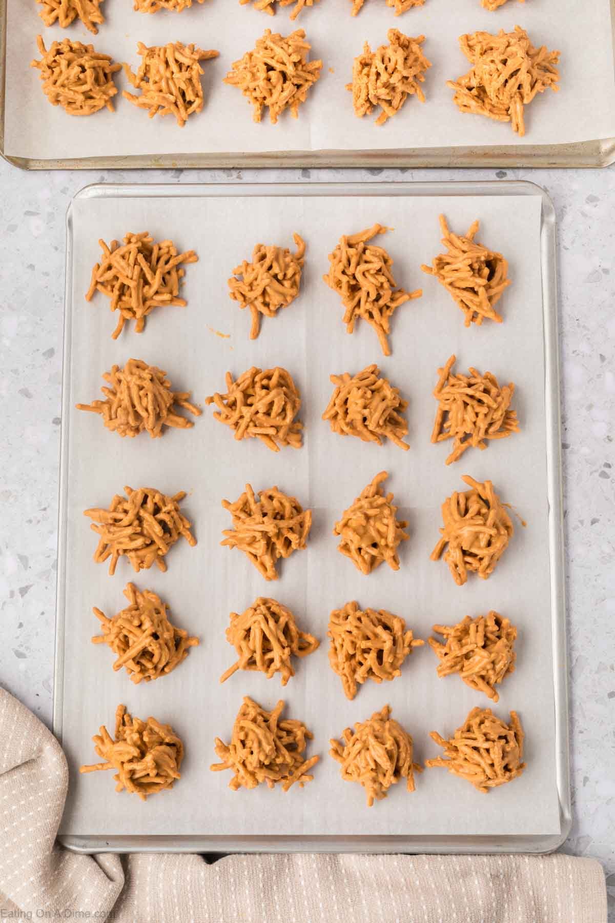 A baking sheet lined with parchment paper holds rows of Haystack Cookiesโeasy no bake cookies made of chow mein noodles coated in a light brown mixture, with more cookies visible on a tray in the background.
