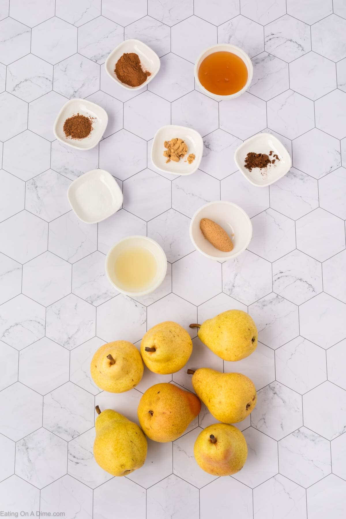 Overhead view of seven yellow pears and eight small bowls containing Pear Butter Recipe ingredients like honey, spices, lemon juice, and brown sugar, all arranged neatly on a white hexagon-patterned surface.