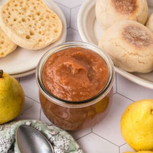 A glass jar filled with homemade pear butter sits on a table, surrounded by fresh pears, a spoon, a patterned cloth, and plates with sliced and whole English muffins—an inviting scene for trying your favorite Pear Butter Recipe.
