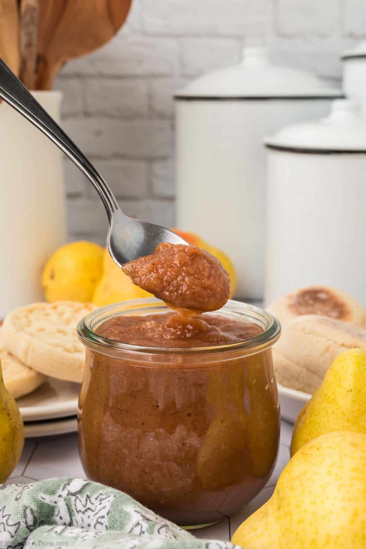A spoon scoops brown pear butter from a glass jar, surrounded by fresh pears, English muffins, and kitchen canisters on a white countertop—perfect inspiration for your next pear butter recipe.