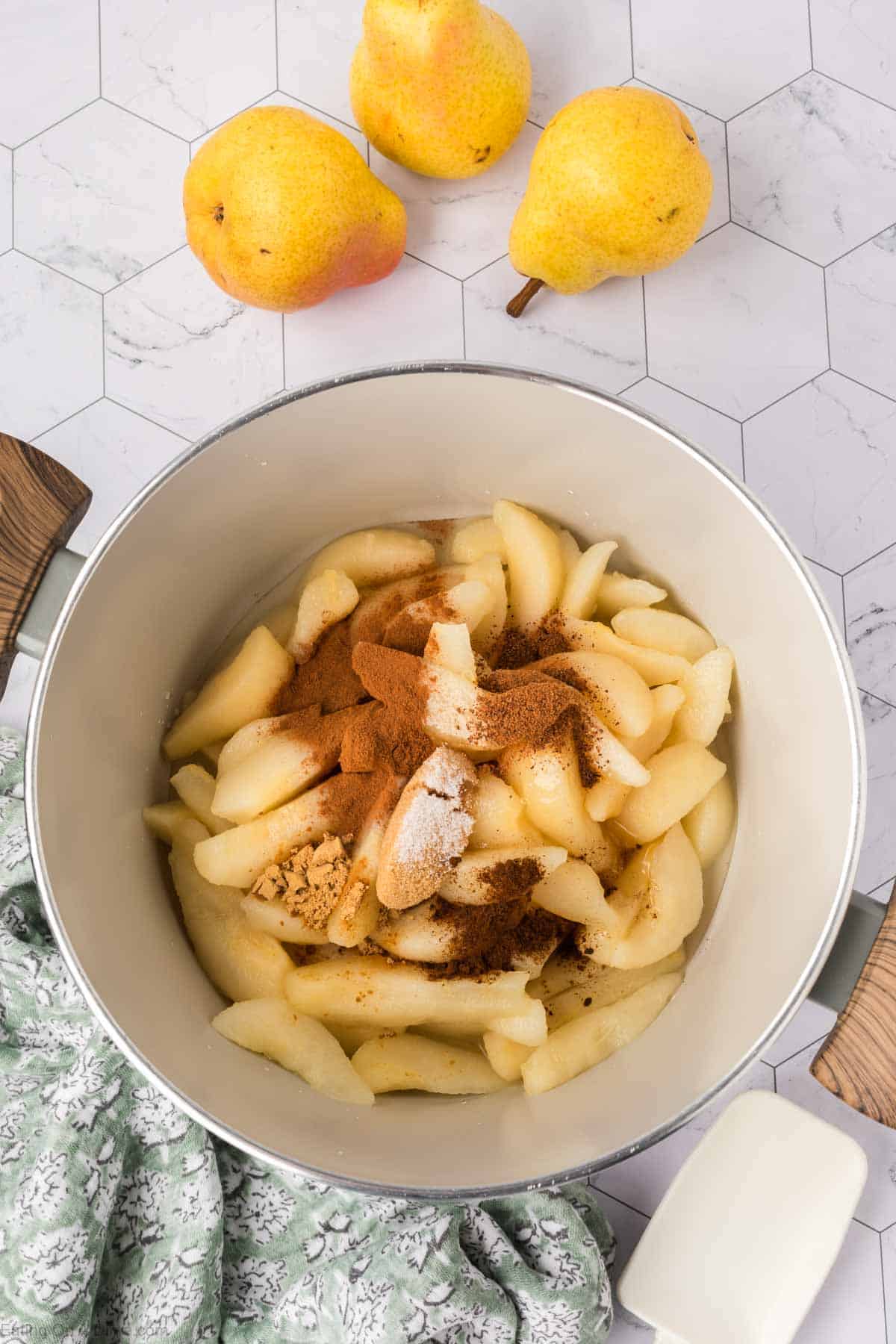 Sliced pears in a pot topped with cinnamon and spices, ready to be cooked for a delicious pear butter recipe. Three whole pears sit on the countertop nearby, along with a green patterned cloth and a white cup.