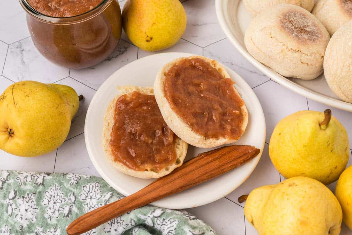 A plate with an English muffin spread with homemade pear butter recipe, surrounded by fresh pears, a jar of pear butter, a wooden spreader, and more English muffins on a white surface.