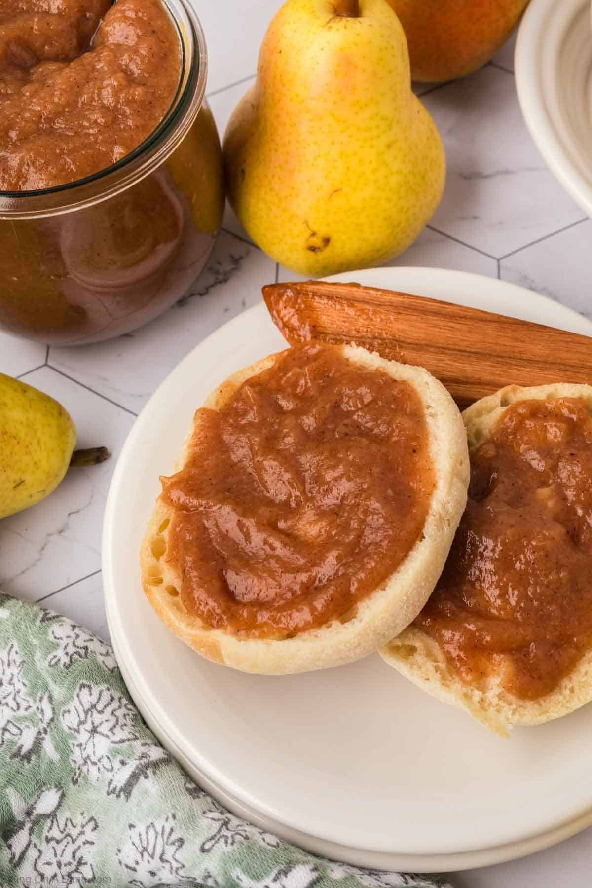 An English muffin spread with homemade pear butter recipe sits on a white plate next to cinnamon sticks, a jar of pear butter, fresh pears, and a patterned cloth on a white surface.