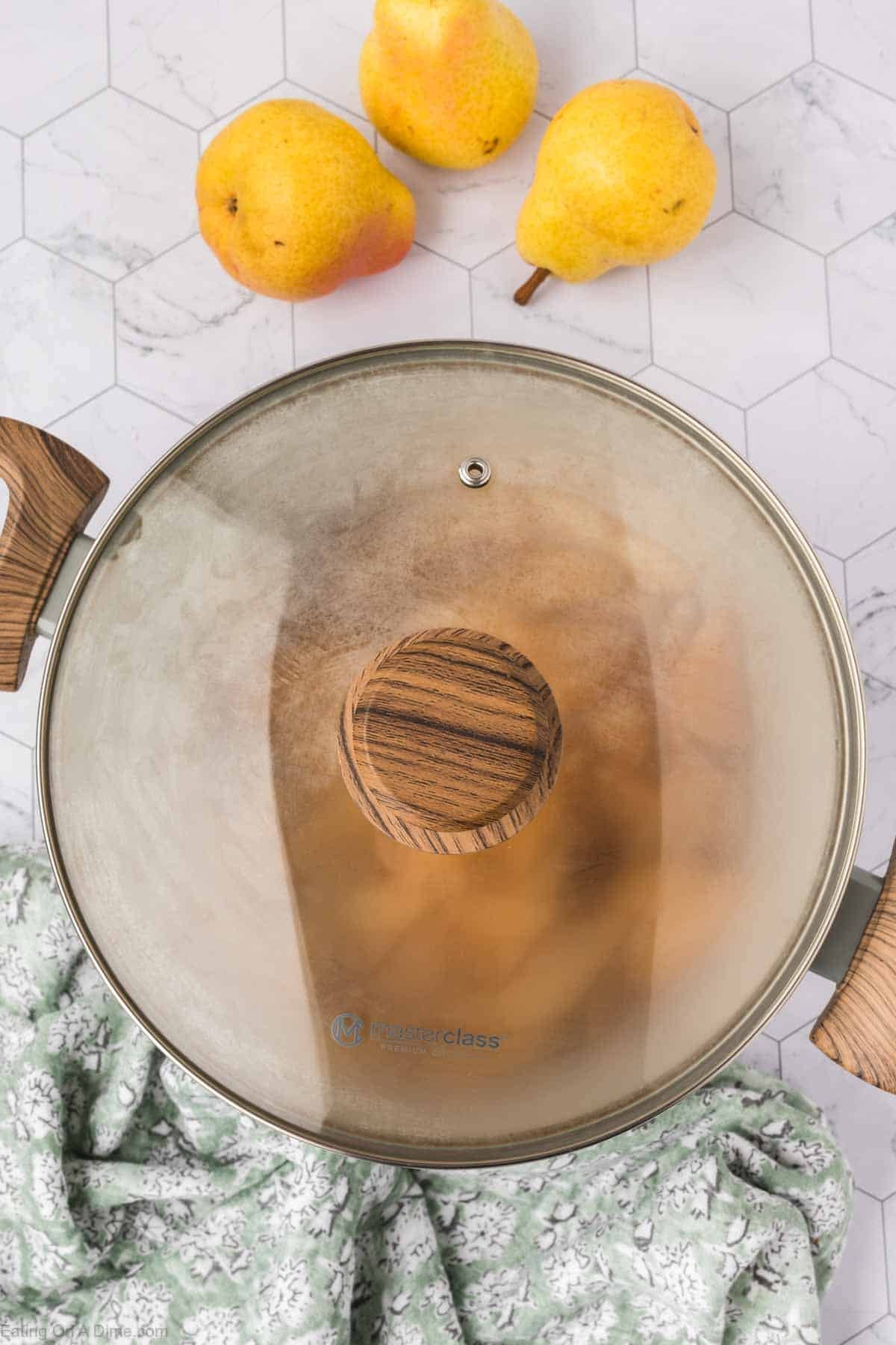 A pot with a wooden handle and glass lid sits on white hexagonal tiles, partially covered by a green patterned cloth; three yellow pears—perfect for your next Pear Butter Recipe—are placed above the pot.
