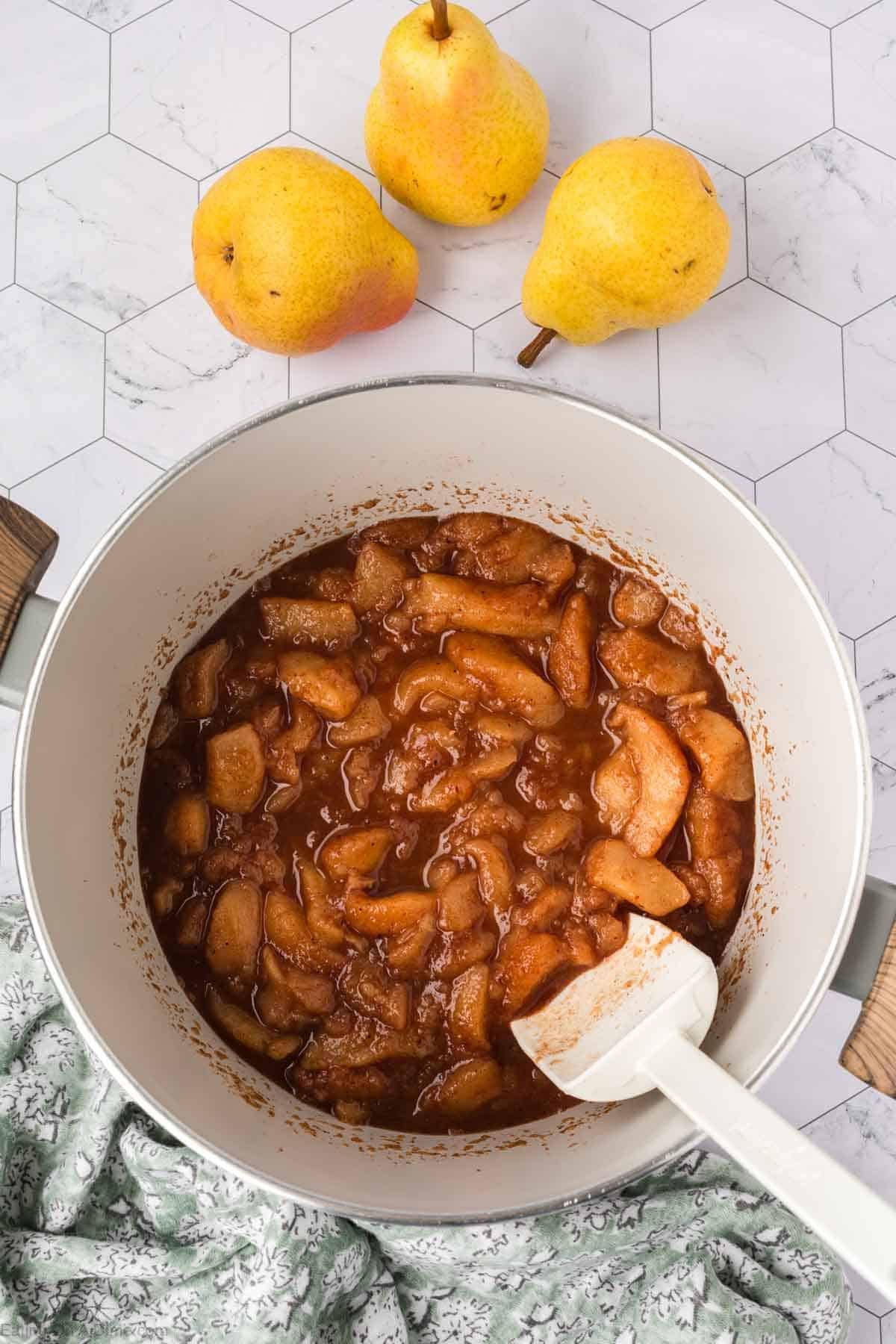 Overhead view of a pot filled with cooked, spiced pear slices for a delicious pear butter recipe. A white spatula rests inside the pot, with three whole yellow pears and a green patterned cloth nearby on a white hexagonal tile surface.