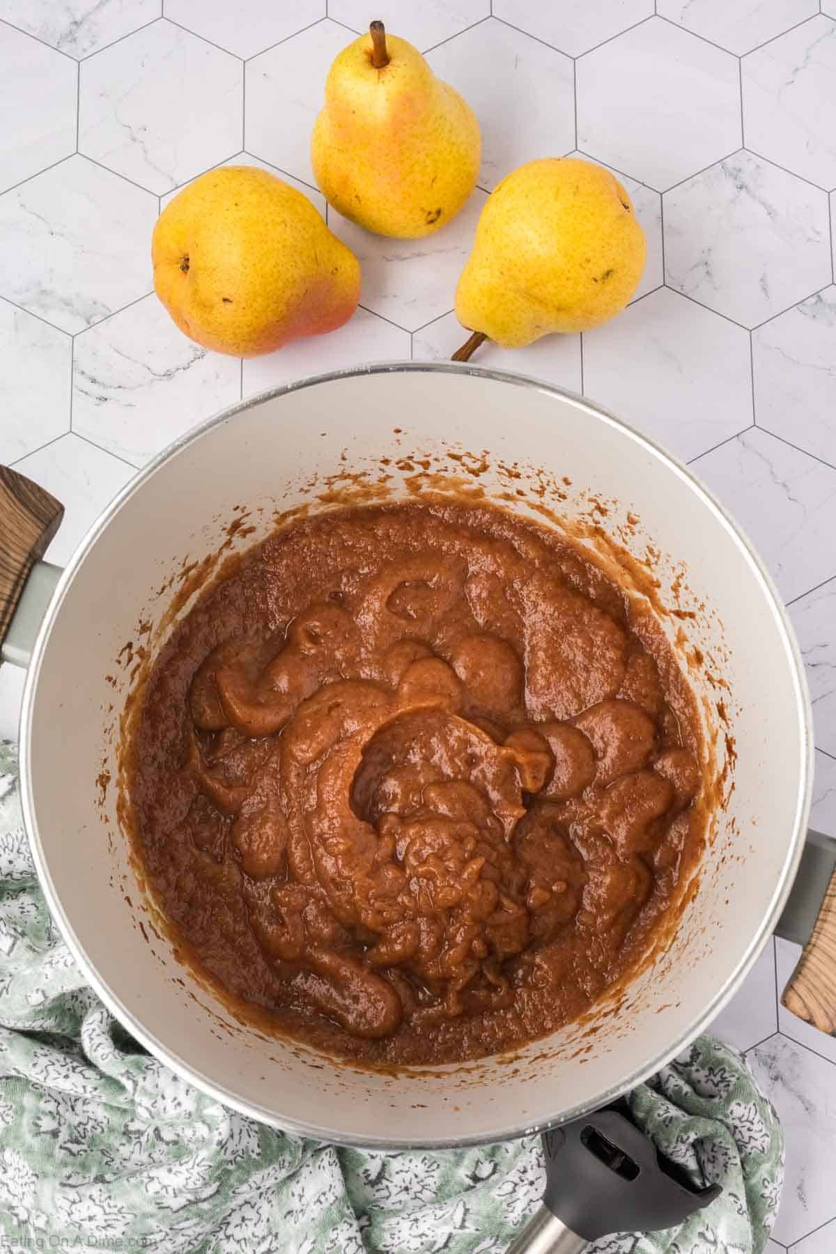 A pot filled with thick brown pear butter sits on a white hexagon-tiled countertop, alongside three whole yellow pears and a green patterned cloth—perfect for trying out your favorite pear butter recipe.