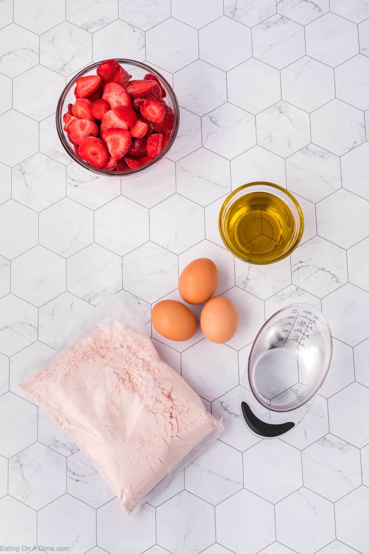 A bowl of sliced strawberries, oil, eggs, water, and a bag of pink cake mix are arranged on a hexagon-tiled surface—everything you need to make a delicious Strawberry Poke Cake.