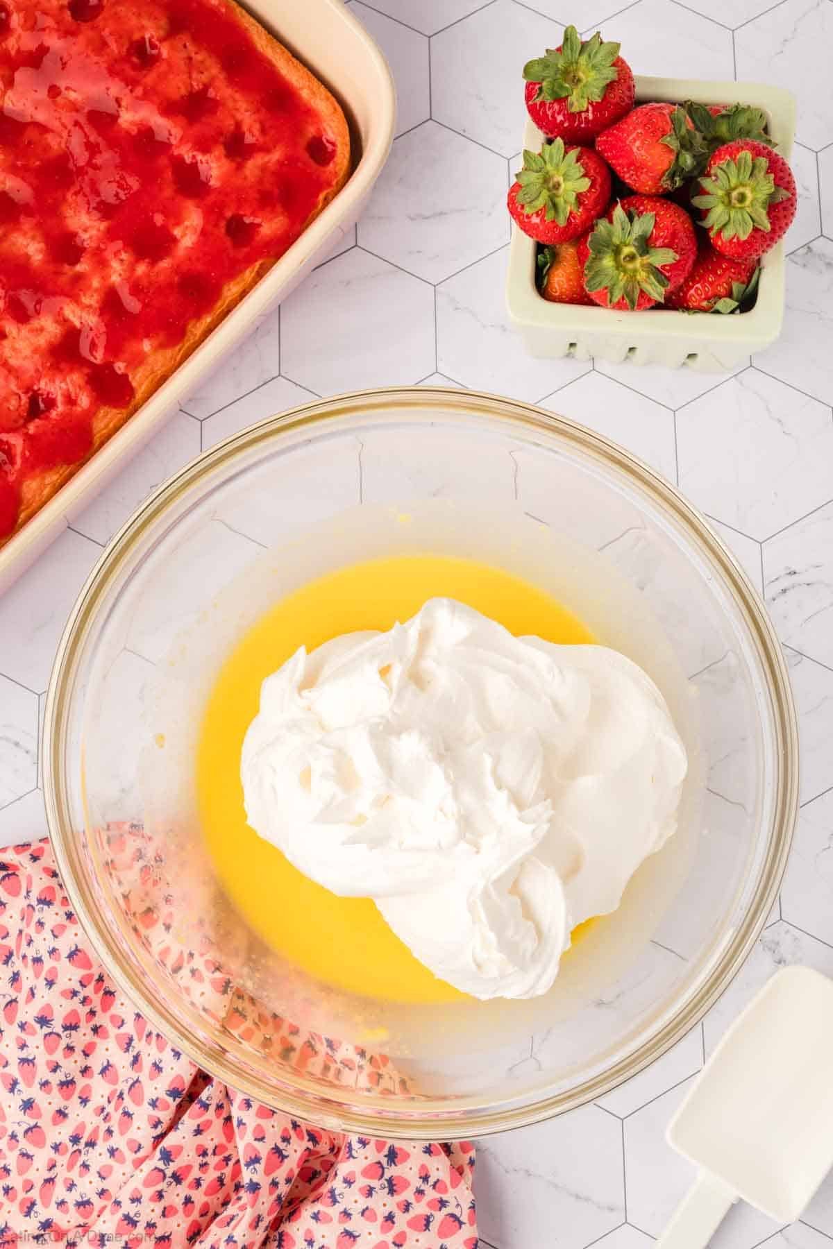 A bowl with whipped topping and yellow pudding mixture sits on a white counter near a carton of strawberries and a baking dish with Strawberry Poke Cake, alongside a pink floral cloth and a white spatula.