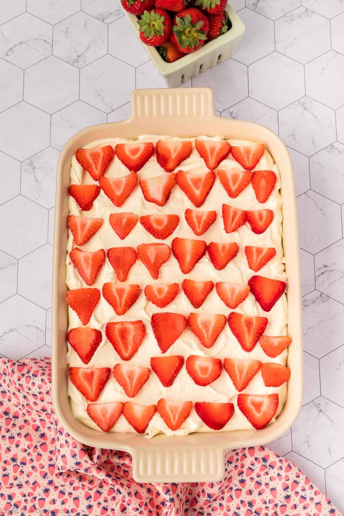 A rectangular baking dish filled with creamy Strawberry Poke Cake topped with rows of sliced strawberries, placed on a hexagonal-tiled surface next to a small container of whole strawberries and a pink patterned cloth.