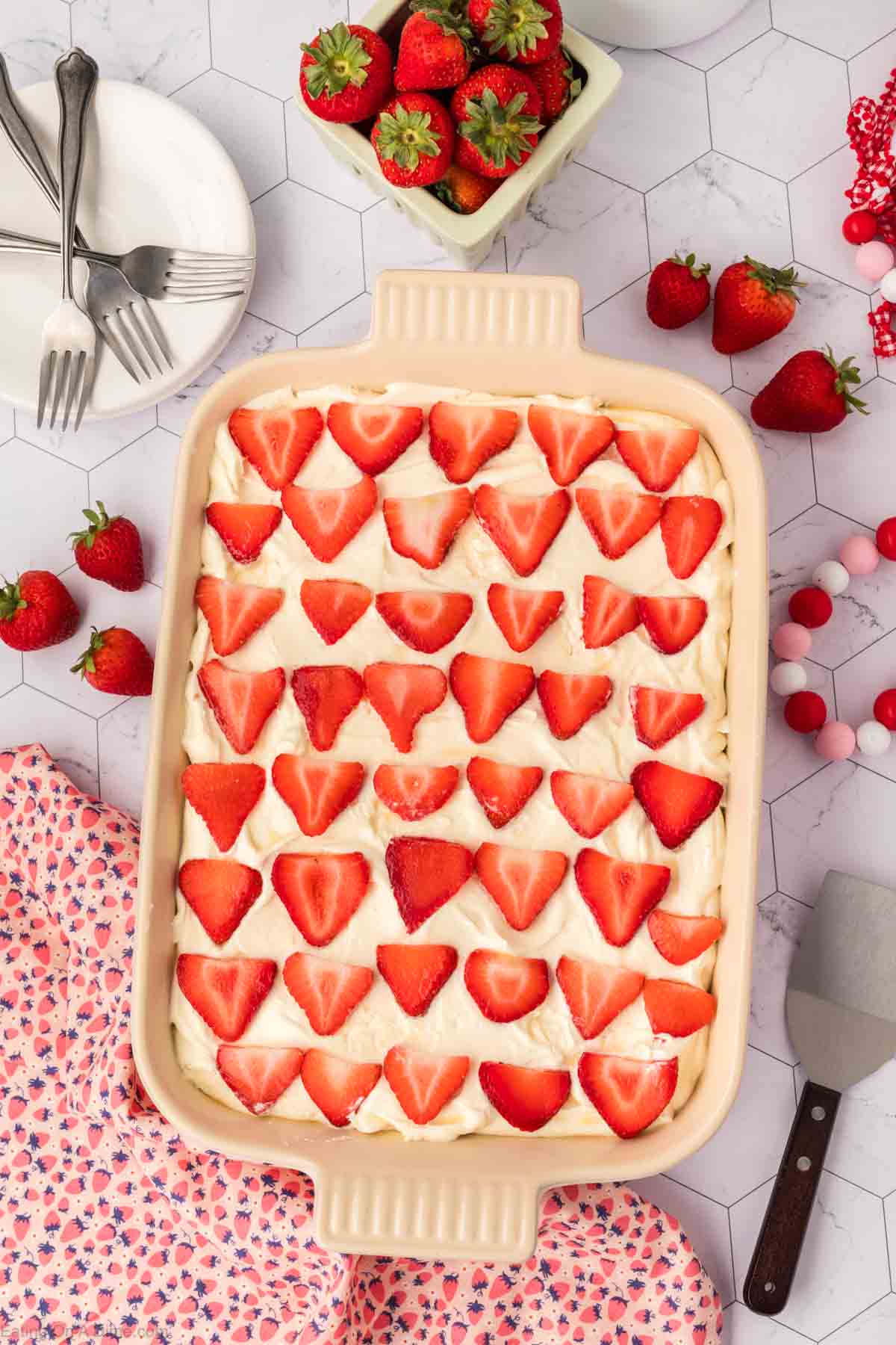 A baking dish filled with creamy Strawberry Poke Cake topped with rows of halved strawberries, surrounded by fresh strawberries, a floral napkin, a spatula, and a plate with forks on a white geometric countertop.