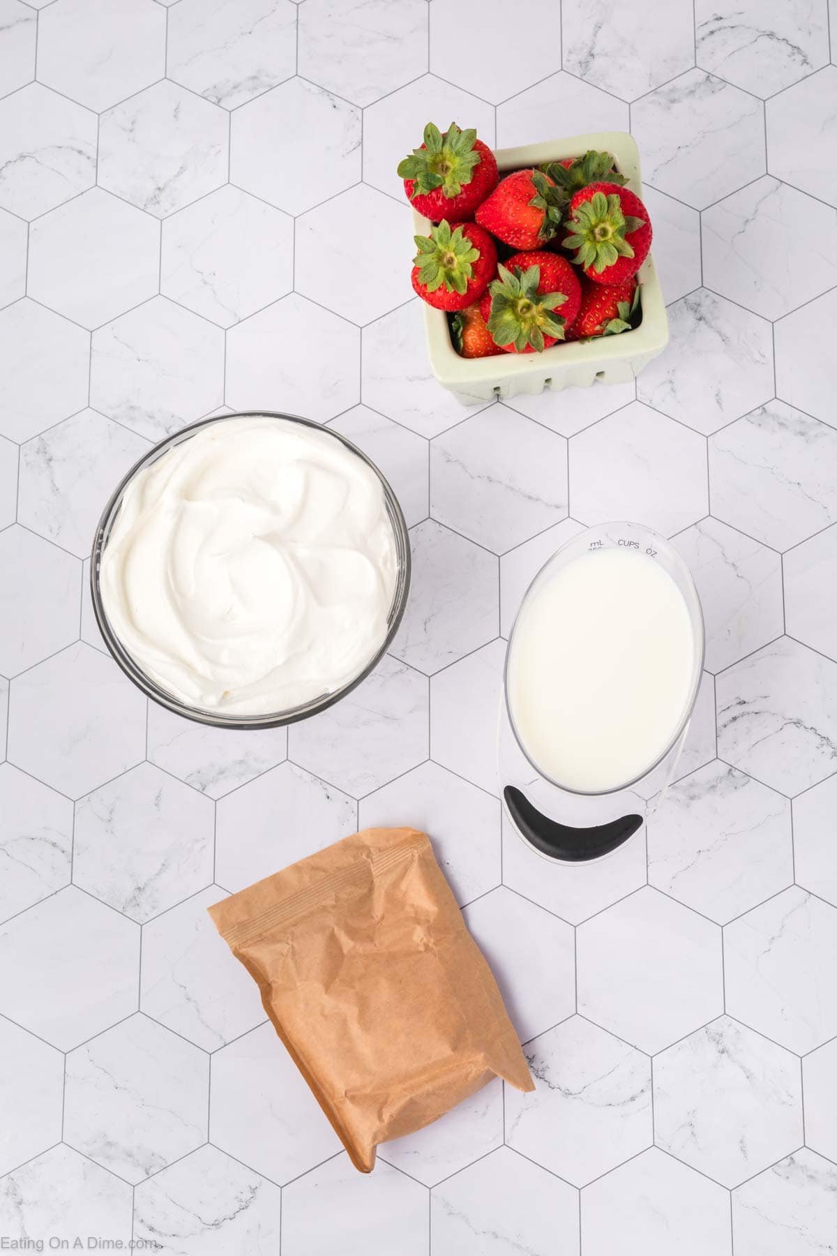 A bowl of whipped cream, a small pitcher of milk, a brown paper packet, and a basket of fresh strawberries arranged on a white hexagonal tile surface—perfect for making a delicious Strawberry Poke Cake.