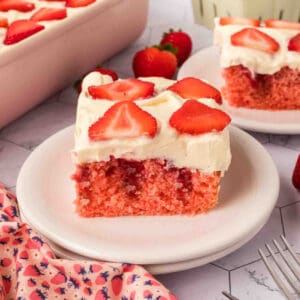 A slice of Strawberry Poke Cake with white frosting and fresh strawberry slices on top sits on a white plate, surrounded by whole strawberries and more cake in the background.