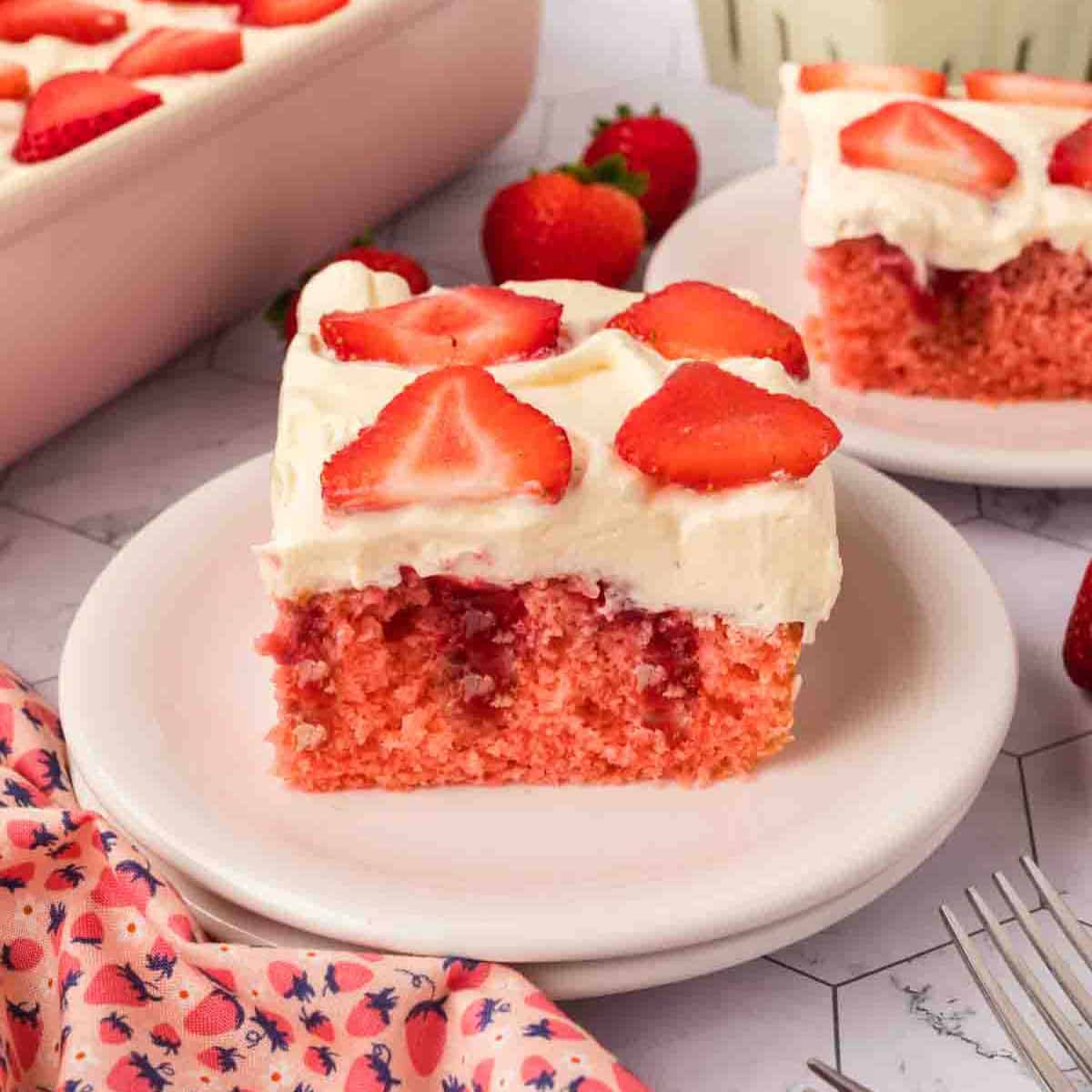 A slice of Strawberry Poke Cake with white frosting and fresh strawberry slices on top sits on a white plate, surrounded by whole strawberries and more cake in the background.