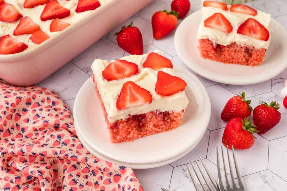 A slice of Strawberry Poke Cake with white frosting and fresh strawberry slices sits on a white plate. More cake, strawberries, a floral napkin, and two forks are nearby on a marble surface.