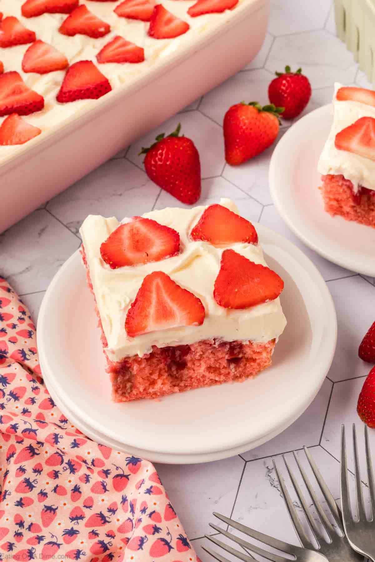 A slice of Strawberry Poke Cake with white frosting and fresh strawberry slices on top, served on a white plate. Whole strawberries and another slice of cake are nearby, with a pink floral cloth and silver forks beside them.