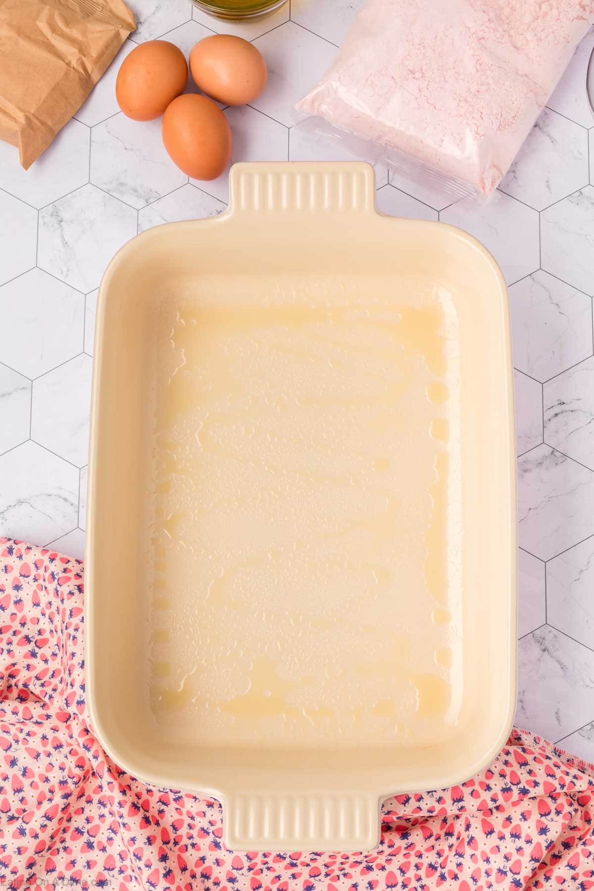 A greased rectangular baking dish sits on a white hexagon-tiled surface, ready to become a delicious Strawberry Poke Cake, surrounded by three eggs, a bag of flour, brown paper, a bottle of oil, and a pink floral cloth.