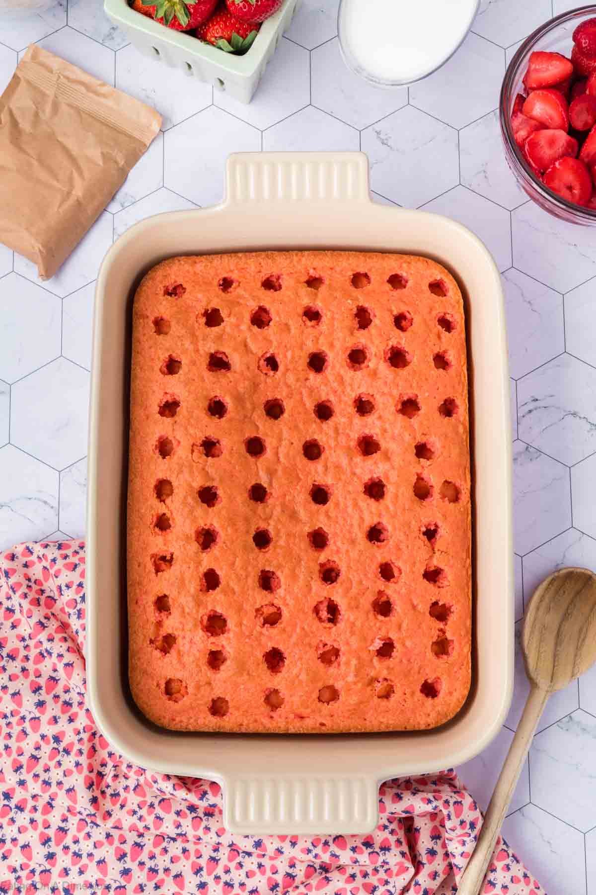 A rectangular Strawberry Poke Cake in a baking dish with evenly spaced holes on top, surrounded by a wooden spoon, a floral cloth, and bowls of strawberries and sugar on a white hexagon-tiled surface.