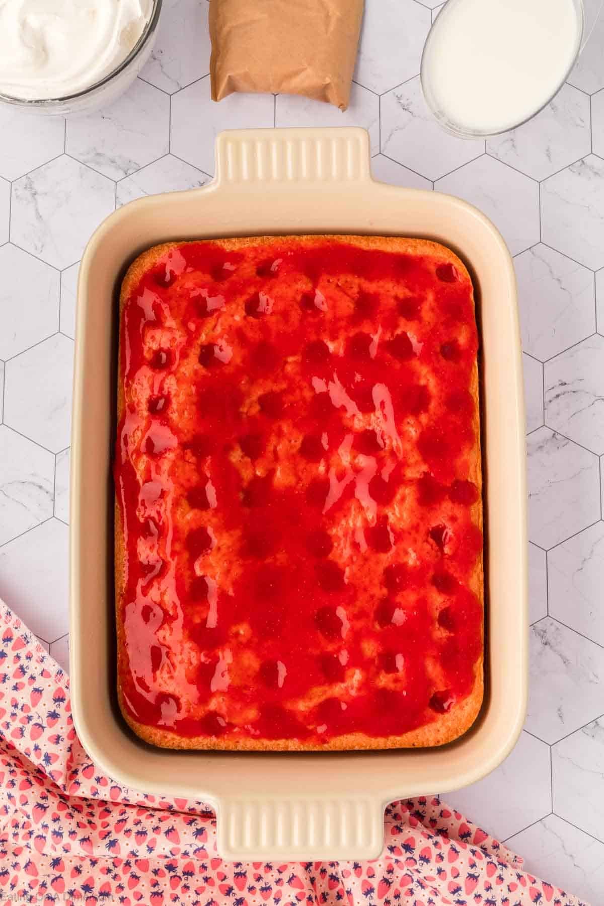 A rectangular baking dish filled with Strawberry Poke Cake topped with a glossy red sauce. Surrounding the dish are a bowl of whipped cream, a brown packet, and a glass of milk on a white, hexagon-tiled surface.