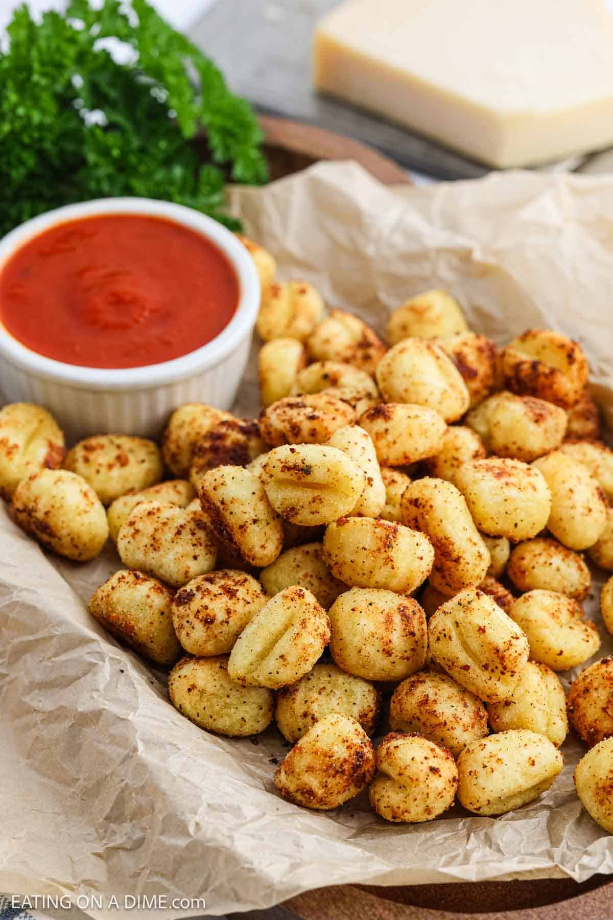 A pile of seasoned, roasted air fryer gnocchi sits on parchment paper with a small dish of marinara sauce and a sprig of parsley on the side. A block of cheese is visible in the background.