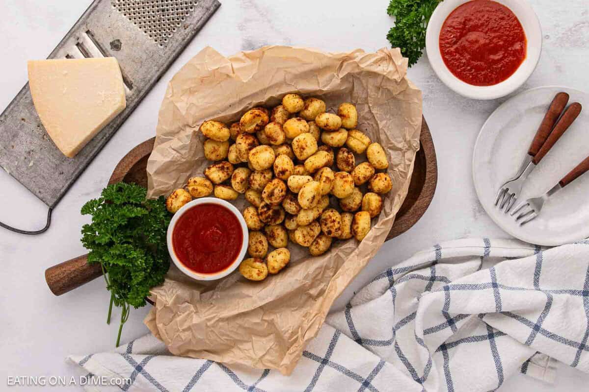 A serving tray lined with parchment paper holds roasted air fryer gnocchi beside a small bowl of marinara sauce, surrounded by parsley, a wedge of cheese, a grater, utensils, and a checkered towel on a white surface.