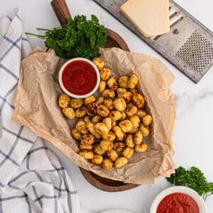 A wooden board lined with parchment paper holds golden, crispy Air Fryer Gnocchi next to a bowl of red marinara sauce. Fresh parsley, a block of cheese, a grater, and a checkered cloth are nearby on a white surface.