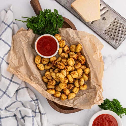 A wooden board lined with parchment paper holds golden, crispy Air Fryer Gnocchi next to a bowl of red marinara sauce. Fresh parsley, a block of cheese, a grater, and a checkered cloth are nearby on a white surface.
