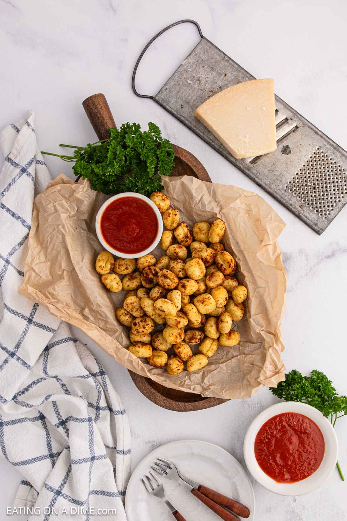 A wooden platter lined with parchment paper holds seasoned, Air Fryer Gnocchi and a small bowl of marinara sauce. Nearby are a block of cheese, a grater, fresh parsley, and a checkered kitchen towel.
